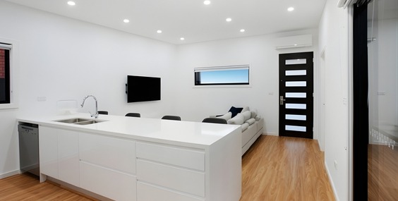 A modern open plan living space with a white kitchen island, couch, television, and black front door, representing factors that influence granny flat approval cost.